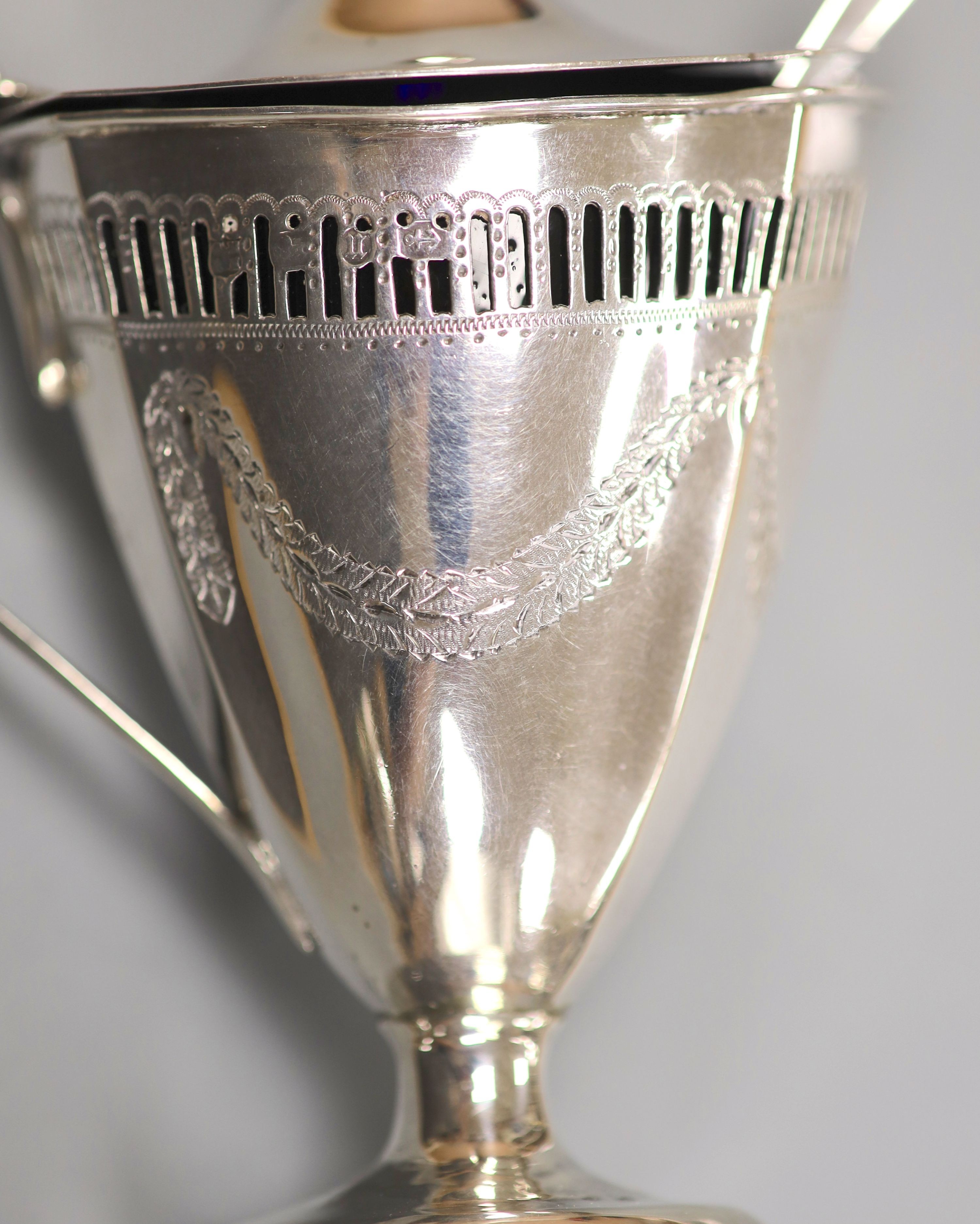 A late Victorian pierced and engraved silver vase shaped pedestal mustard, Nathan & Hayes, Birmingham, 1894, with blue glass liner and an earlier Georgian mustard ladle, height 13.2cm, 147 grams.